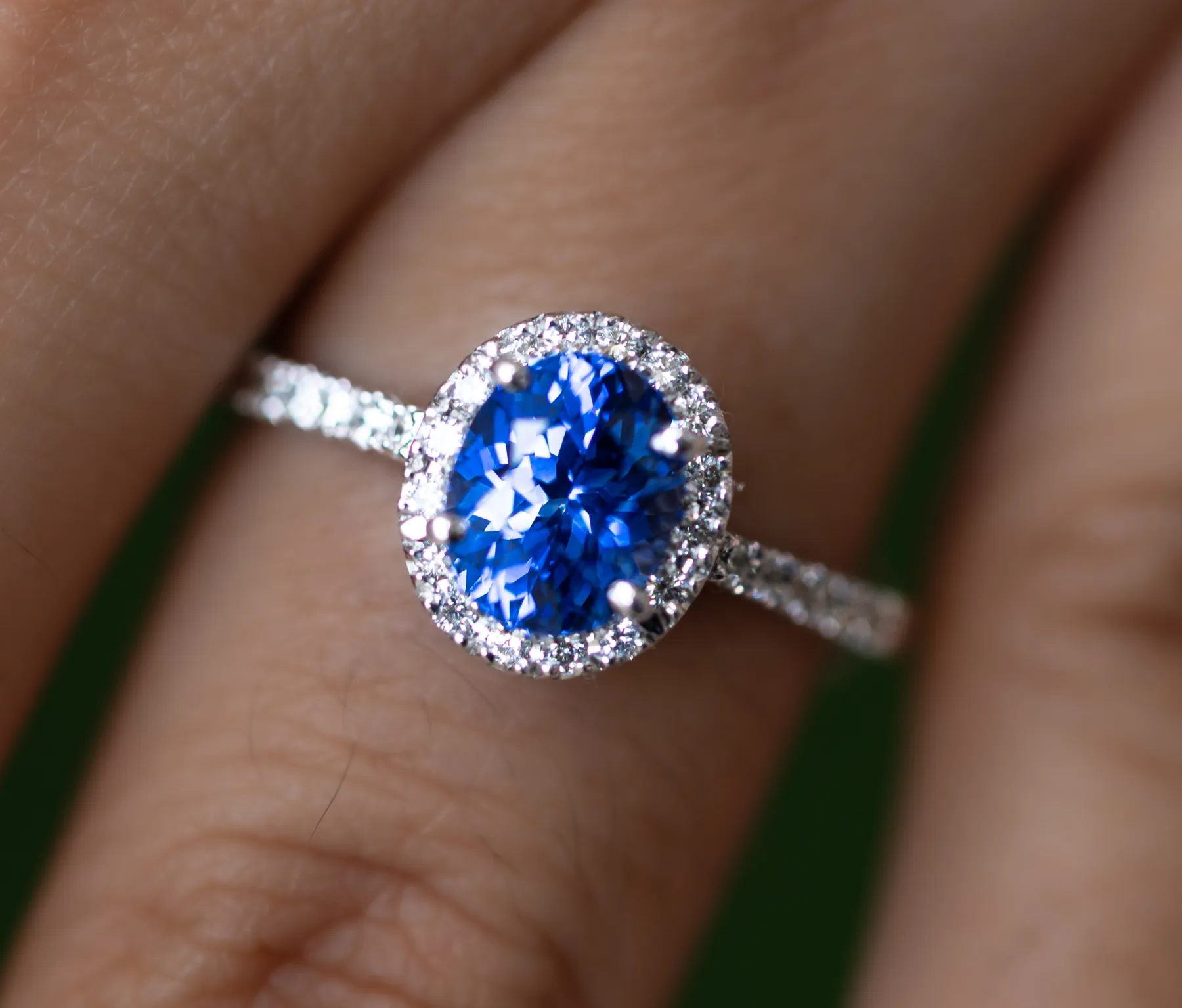 Oval blue Ceylon sapphire ring with a diamond halo and pavé band, worn on a finger