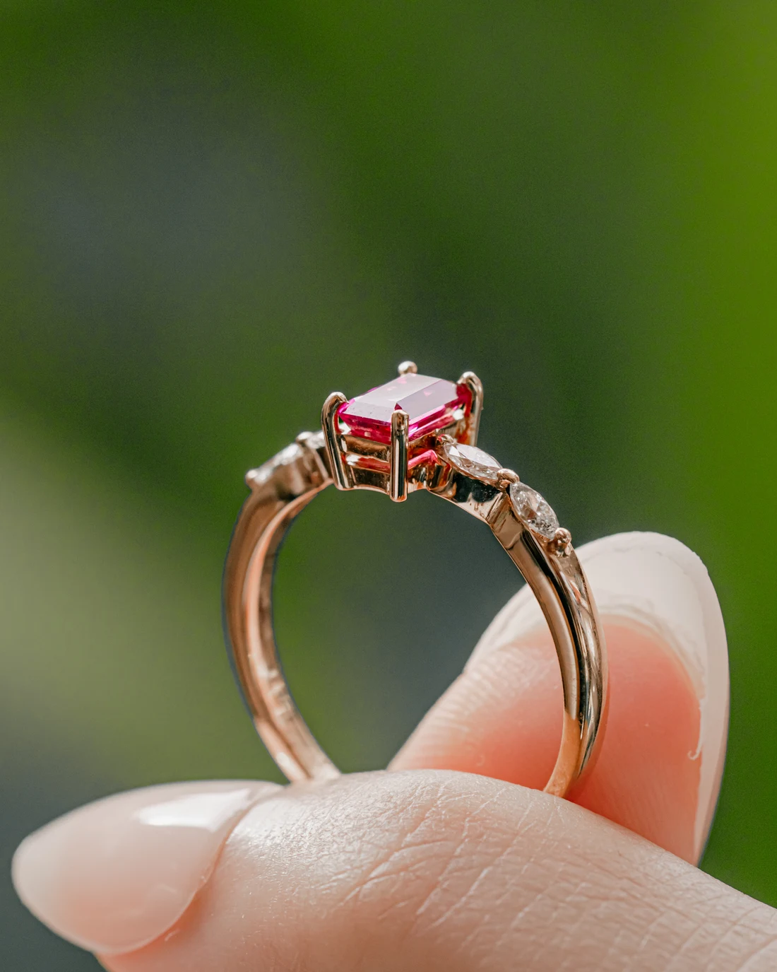 Ruby emerald-cut ring with marquise diamonds in rose gold, held against green leaves